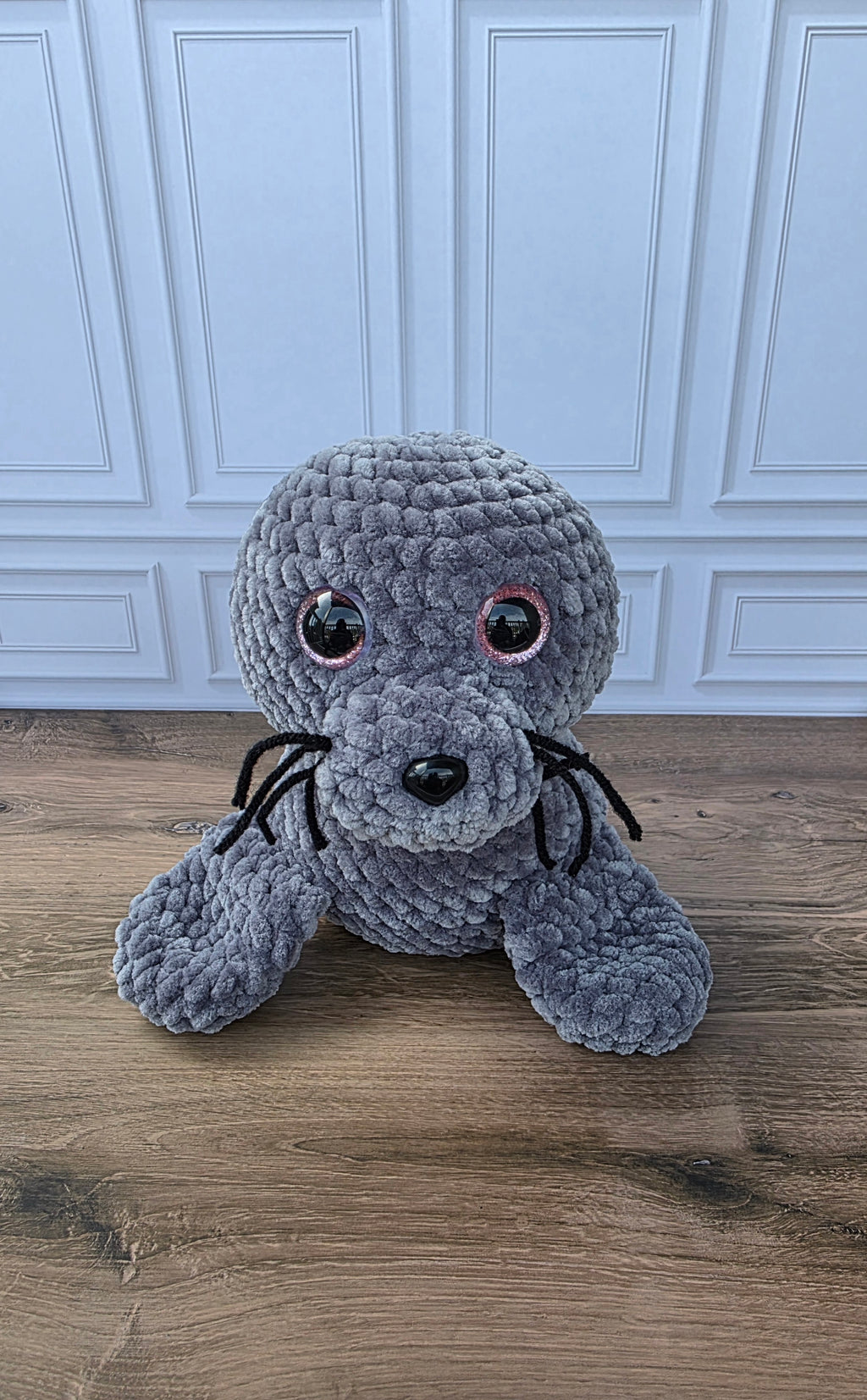 A gray crocheted seal plush toy with large black pupils encircled in pink color, and black whiskers, sitting on a wooden floor in front of a white paneled wall