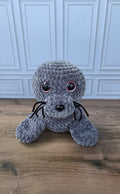 A gray crocheted seal plush toy with large black pupils encircled in pink color, and black whiskers, sitting on a wooden floor in front of a white paneled wall
