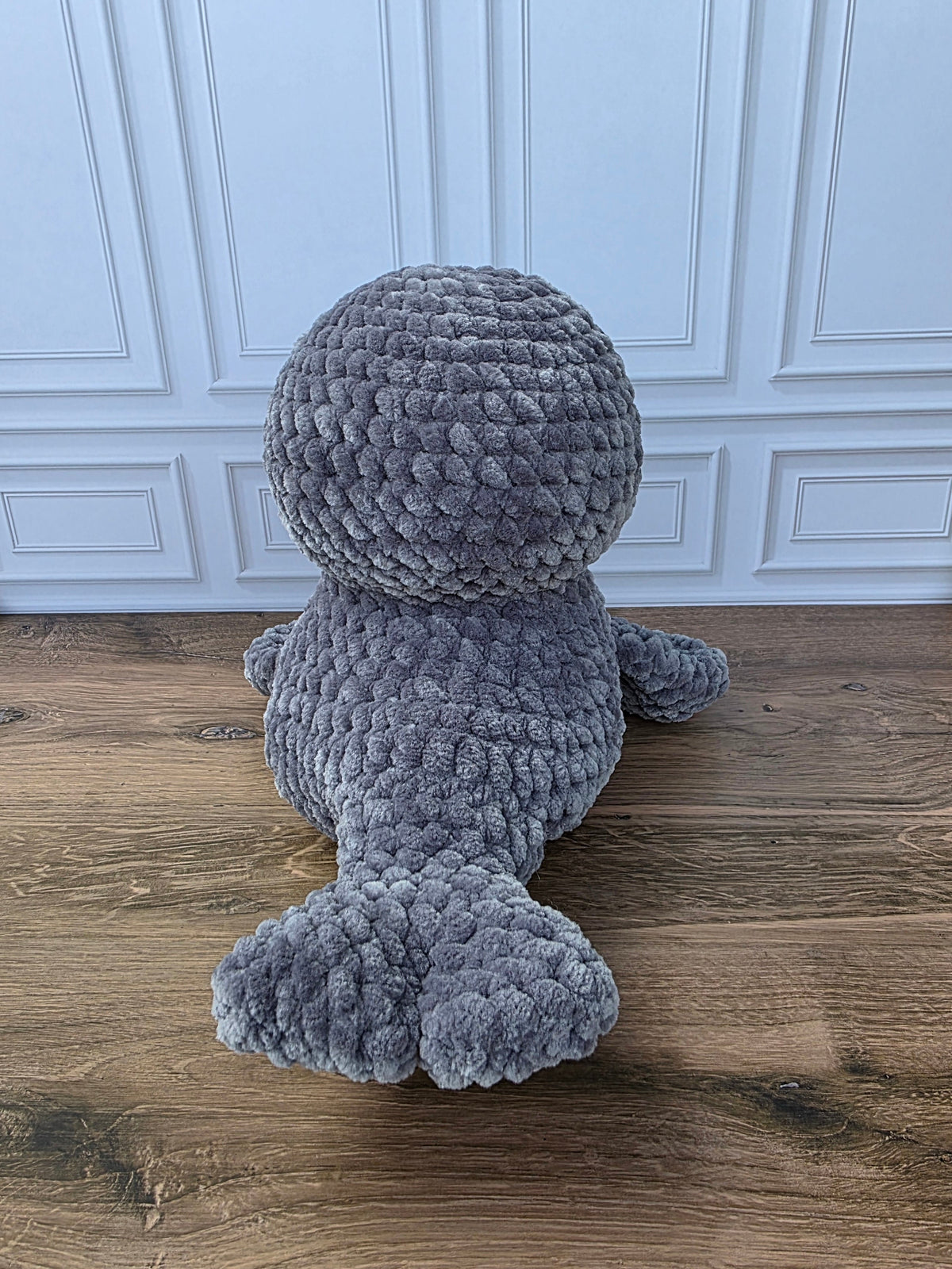 A gray textured plush seal toy shown from the back, sitting on a wooden floor in front of a white paneled wall.