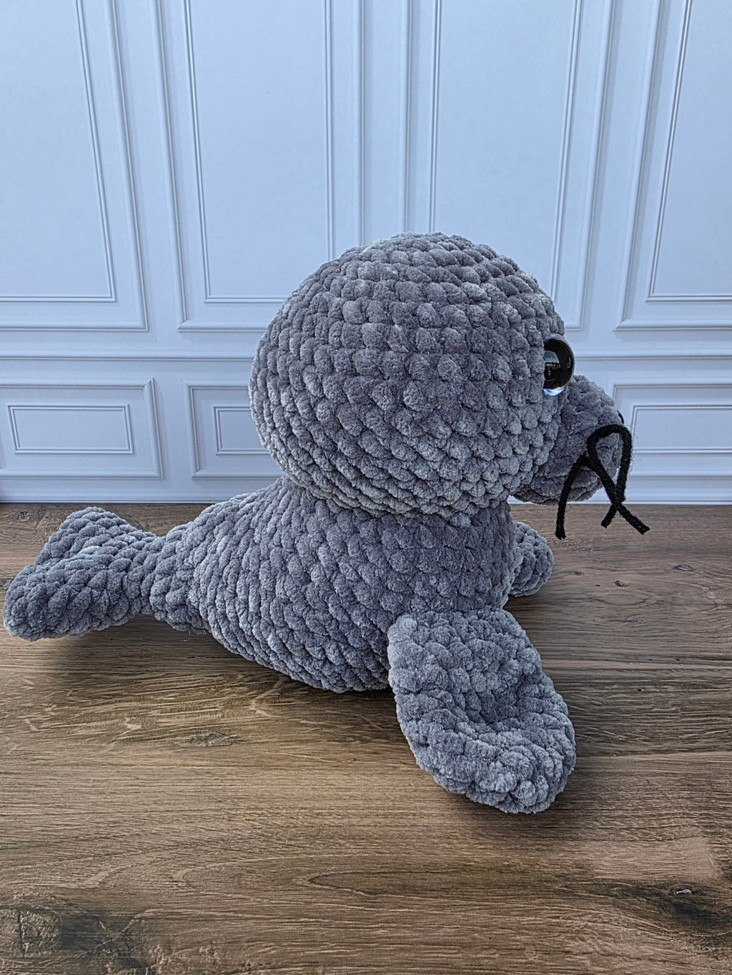 A gray crocheted seal plush toy lying on a wooden floor in front of a white paneled wall.